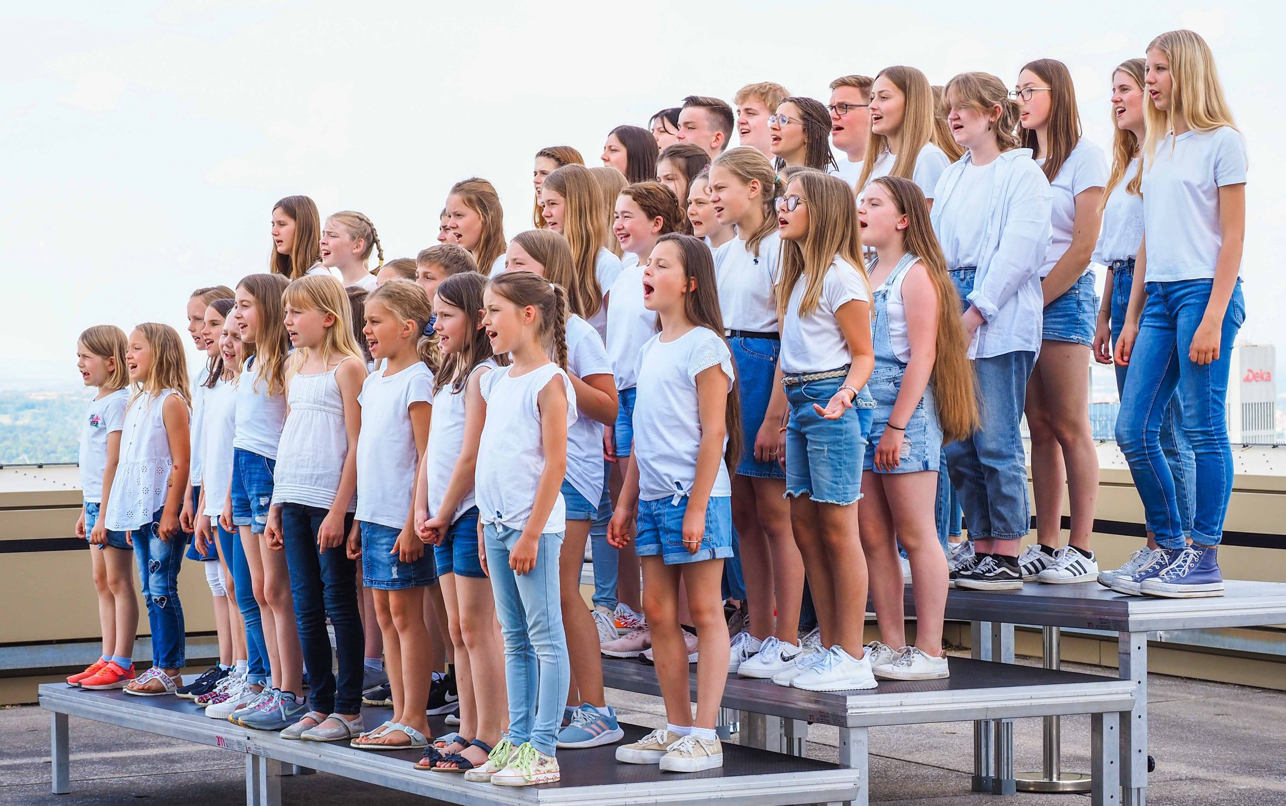 circa 40 singende Kinder auf Podesten stehend, in weißen Oberteilen und Jeanshosen