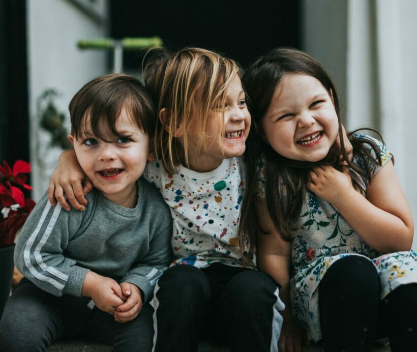 Drei Kinder nebeneinander sitzend lachend auf einer Treppe. Das mittlere Kind umarmt die anderen beiden Kinder.