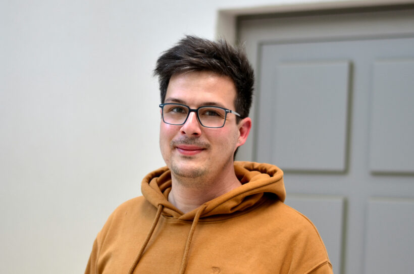 junger Mann mit kurzen dunklen Haaren, dunkler Brille und einem senfgelben Pullover