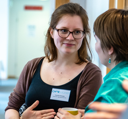 Zwei Personen im Gespräch. Eine mit mittellangen braunen Haaren und einer Hornbrille, eine andere Person seitlich von hinten zu sehen mit kurzen braunen Haaren und einem türkisen Ohrring.
