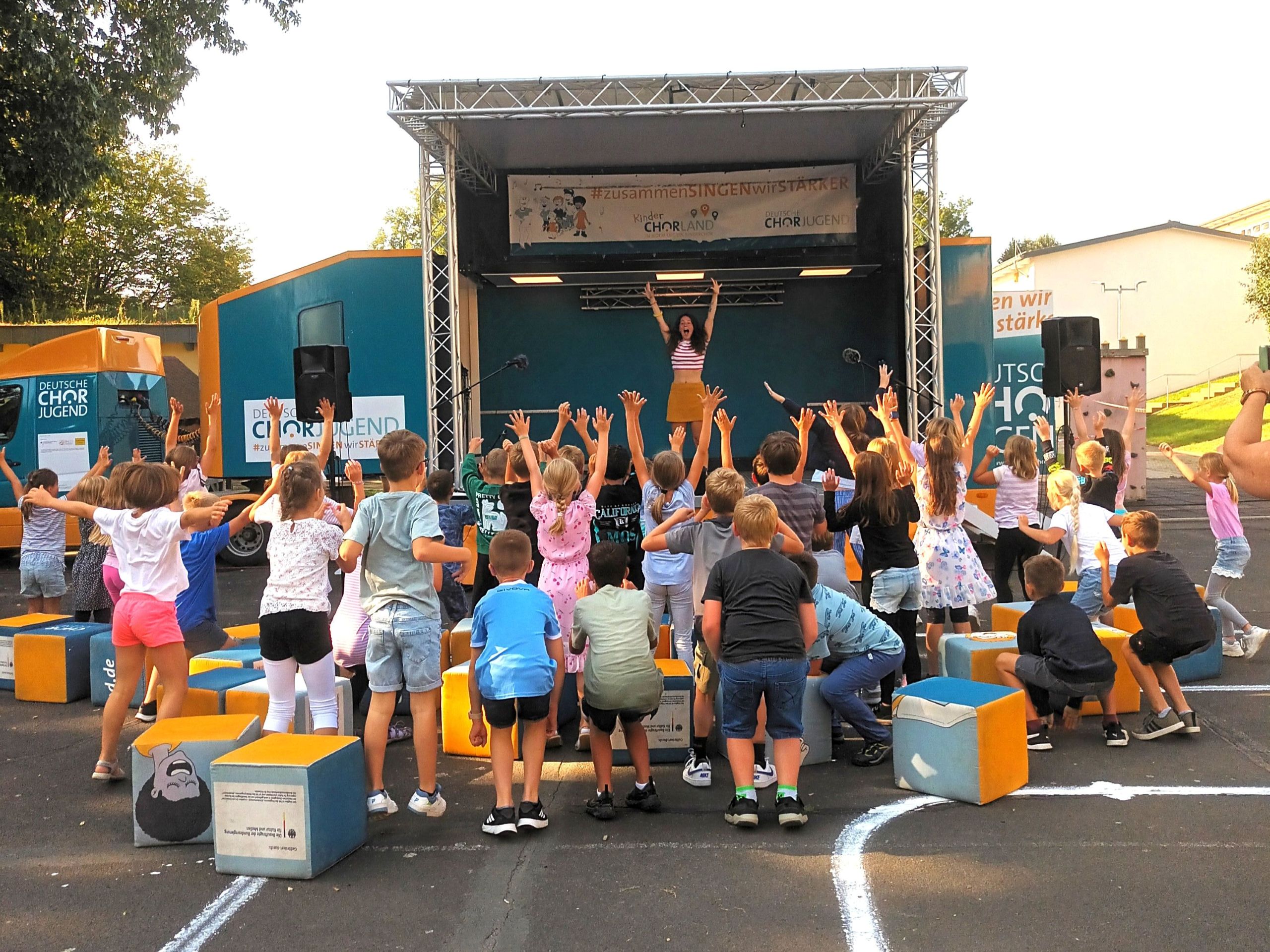 Eine Musikpädagogin steht auf dem SingBus und streckt die Arme nach oben. 40 zu ihr gewandte Kinder tun es ihr gleich. Die Stimmung ist gut. 