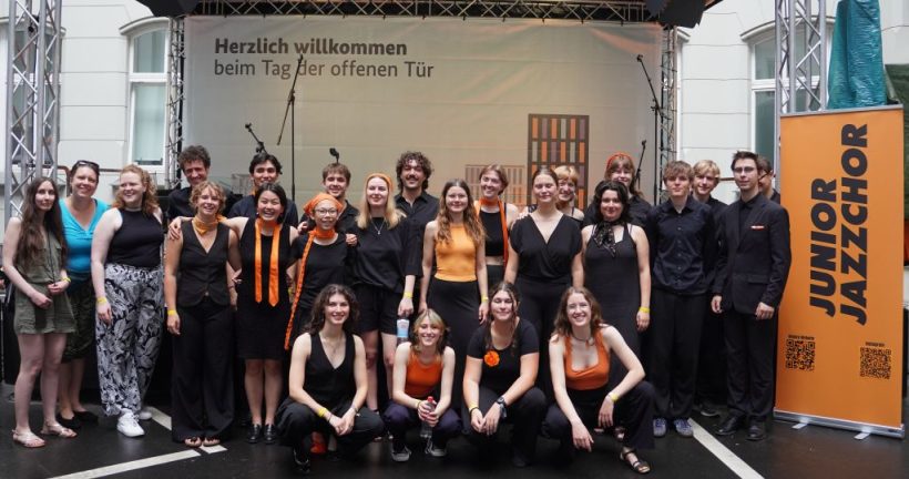 Junge Chorsänger:innen in schwarzer und orangefarbener Kleidung vor einer Bühne.