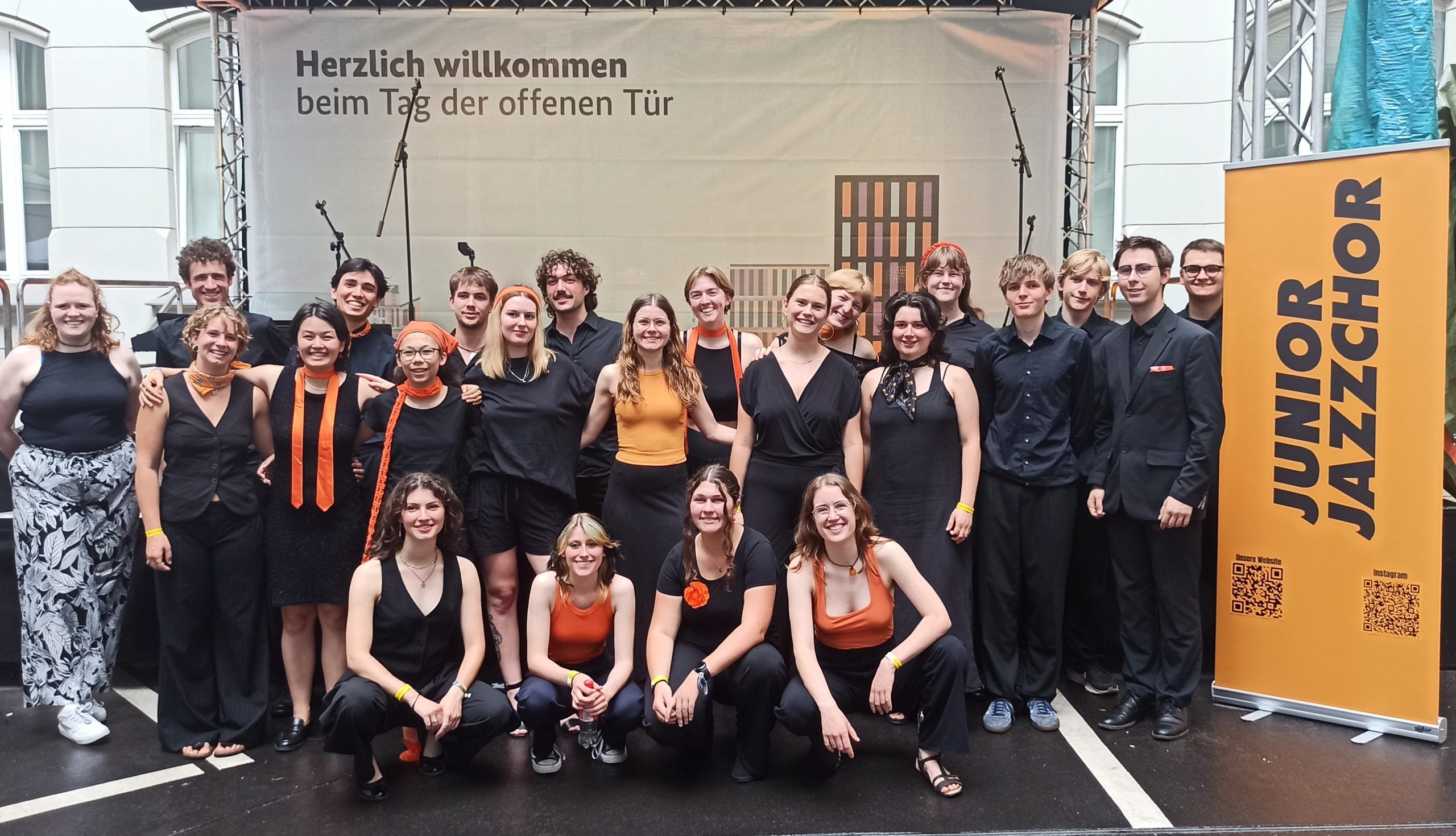 Junge Chorsänger:innen in schwarzer und orangefarbener Kleidung vor einer Bühne.