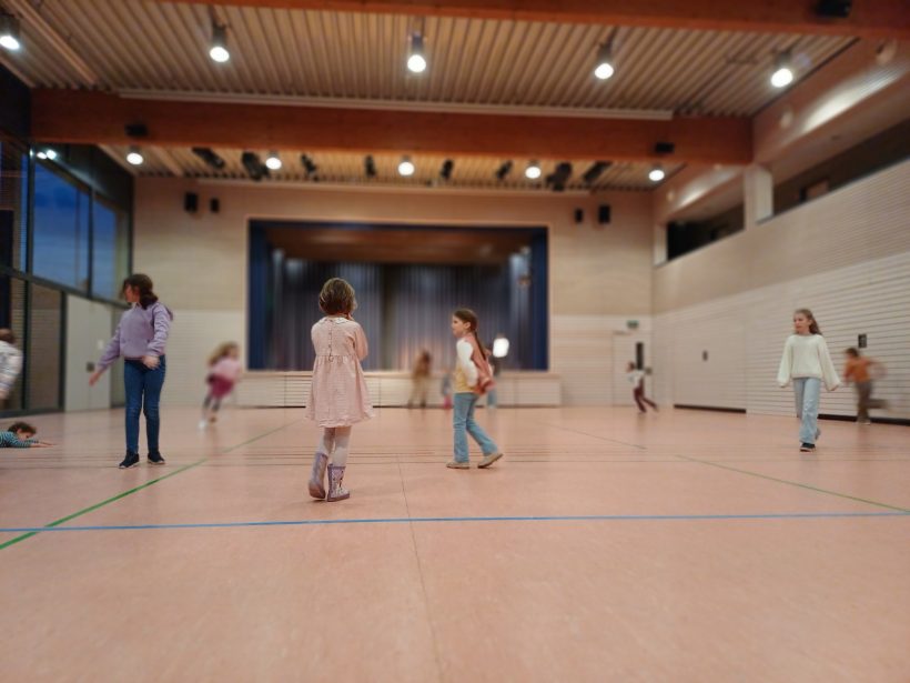 Eine Turnhalle, in der einzelne Kinder hin und her rennen. Ein Kind in rosa Kleid steht alleine in der Mitte und blickt in den Raum.