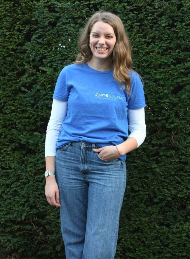 Person mit langen welligen Haaren und blauem T-Shirt mit Chorjugend-Schriftzug
