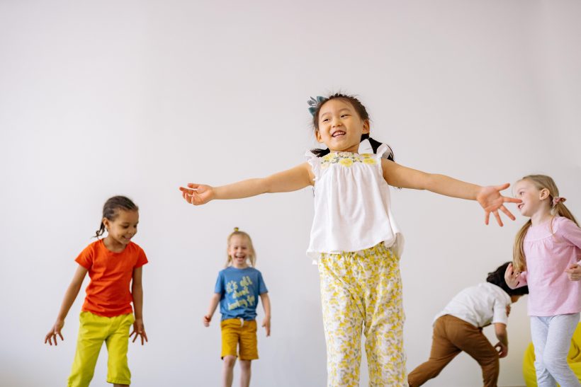 Fünf Kinder vor weißem Hintergrund in Bewegung und bunten Kleidern. Das Kind am vordesten im Bild springt mit ausgestreckten Armen in die Luft.